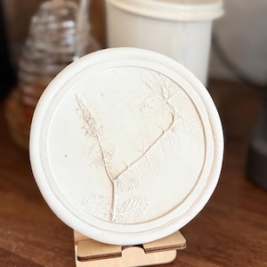 May include: A round, off-white ceramic coaster with a raised botanical design of leaves and a stem. The coaster is displayed on a small wooden stand. Other items in the background include a honey jar and a white container.