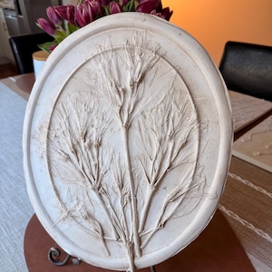 Könnte beinhalten: Ovale, cremefarbene Keramik-Wandkunst mit einem erhabenen botanischen Relief. Das Kunstwerk zeigt einen zentralen Stiel mit mehreren Ästen und Blattdetails. Das Stück wird auf einem dekorativen Metallständer präsentiert.