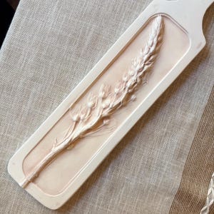 Pode incluir: Bandeja decorativa retangular esbranquiçada com um relevo em relevo de uma espiga de trigo. A espiga de trigo é de cor bege claro e está centrada sobre um fundo bege ligeiramente mais escuro. A bandeja tem um acabamento liso e mate.