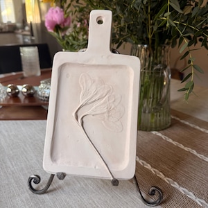 Puede incluir: Una bandeja decorativa rectangular blanca con asa y diseño floral en relieve. La bandeja se exhibe sobre un soporte de hierro forjado. El fondo incluye un jarrón con flores y una mesa.