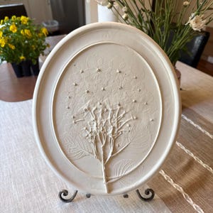 Pode incluir: Arte de parede oval em cerâmica branca com um relevo em relevo de uma planta de hortênsia. A obra de arte é montada em um suporte decorativo de metal preto. O design da hortênsia inclui folhas e cachos de flores detalhados.