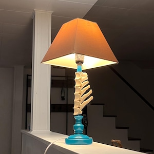 Può includere: Una lampada da tavolo unica con paralume marrone e base turchese. La lampada presenta un design a forma di osso sullo stelo, aggiungendo un tocco distintivo. La lampada è collegata.