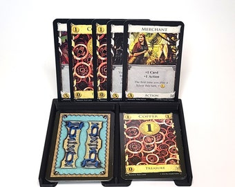 Puede incluir: Primer plano de una configuración de juego de mesa Dominion con cartas en un soporte de madera. Las cartas incluyen "Mercader", "Cobre" y "Provincia".