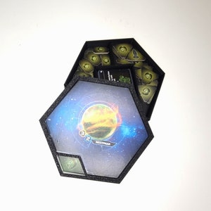 Peut inclure: Une boîte de jeu hexagonale avec un insert en mousse noire et un design sur le thème de l'espace. Le couvercle est ouvert, révélant un compartiment rempli de petits jetons verts. La boîte présente une planète avec une atmosphère bleue et orange et le texte "Westmar" imprimé dessus.