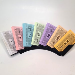 May include: A set of Monopoly game money in denominations of £1, £5, £10, £20, £50, £100, and £500. The money is arranged in a fan shape and is held in a black plastic holder.