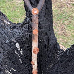 Op de afbeelding: Een handgemaakte houten wandelstok met een koperen handvat en decoratieve koperdraadspiralen. Het handvat is omwikkeld met koperdraad en in het midden is een helder kristal geplaatst. De stok staat tegen een donkere, getextureerde achtergrond.