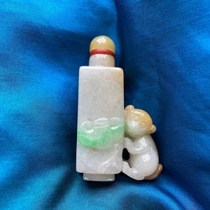 May include: A white jade snuff bottle with a green jade carving of a leaf and a small, white jade monkey figurine attached to the side. The bottle has a red and yellow stopper.