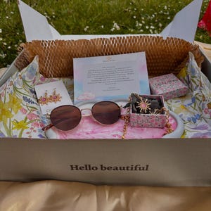 May include: A gift box with the words "Hello beautiful" printed on the front. Inside, a pair of sunglasses, earrings, and a necklace are displayed on a pink tray. The box also contains a small floral-patterned box and a card.