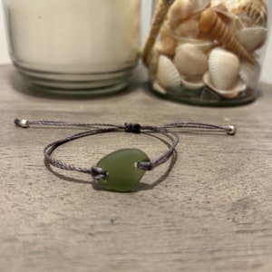以下が含まれることがあります： グレーのコードブレスレットに、緑色のシーグラスのチャームが付いています。チャームはハート型で、中央に小さな穴が開いています。