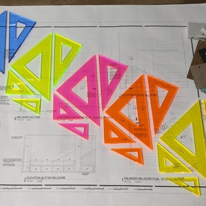 May include: A set of colorful plastic drafting triangles in blue, yellow, pink, and orange on a blueprint. The triangles are arranged in a cascading pattern, with the largest triangle at the bottom and the smallest triangle at the top.
