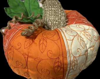 Può includere: Una zucca arancione e bianca fatta a mano con dettagli ricamati. La zucca è fatta di tessuto e ha uno stelo di juta.