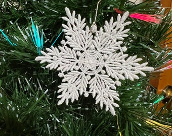 May include: A white snowflake Christmas ornament with a delicate, lacy design hangs on a green artificial Christmas tree with colorful lights.