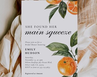 May include: A white bridal shower invitation with a green leafy branch and orange fruit illustration. The text reads "She found her main squeeze" and includes details about the event, including the bride's name, date, time, location, and RSVP information. The invitation also states that the couple is registered at Amazon.
