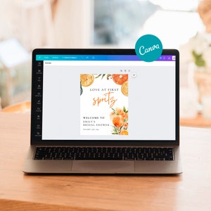 May include: A laptop displays a bridal shower invitation design. The invitation features the text "Love at First Spritz" with orange slice illustrations and floral accents. The design includes the text "Welcome to Emily's Bridal Shower" and the date.