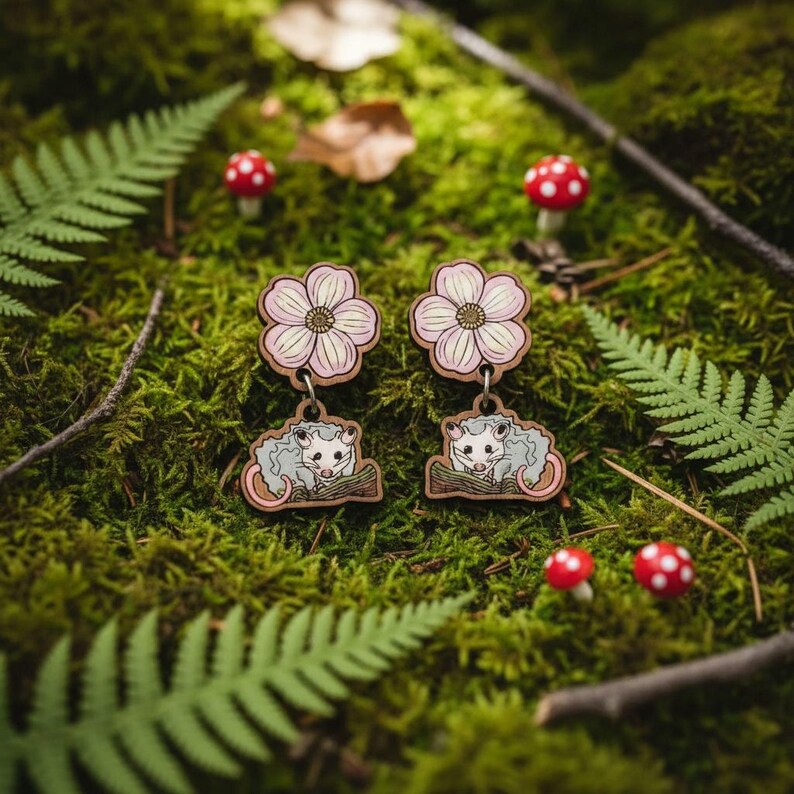 A pair of earrings featuring cartoon opossum characters, surrounded by ferns, mushrooms, and other natural elements, creating a whimsical and nature-inspired scene.