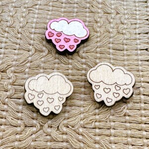 May include: Three wooden cloud ornaments with pink hearts. The top cloud is painted pink with glitter. The other two clouds have hearts cut out of them.