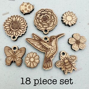 May include: A collection of 18 wooden charms featuring laser-engraved designs including a hummingbird, a butterfly, a bee, a ladybug, and various flower shapes. Each piece has a small hole for hanging, and the text 18 piece set is visible.