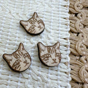 May include: Three wooden cat face buttons with a light brown color and a detailed carved design. The buttons are on a white crocheted surface.