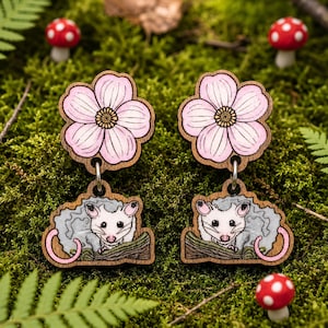 A pair of wooden earrings with a pink flower design and a cartoon opossum illustration, set against a background of green moss and red mushrooms.