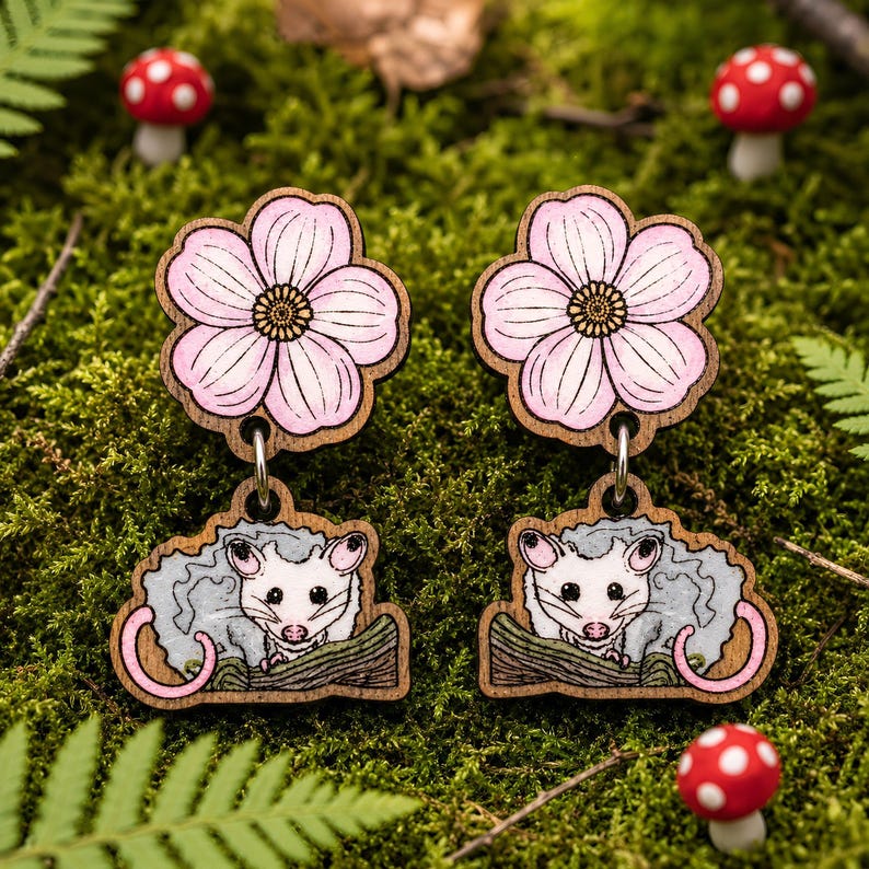 A pair of wooden earrings with a pink flower design and a cartoon opossum illustration, set against a background of green moss and red mushrooms.
