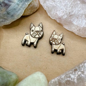 May include: Two wooden French bulldog figurines, one facing left and one facing right. The figurines are light brown with dark brown details.