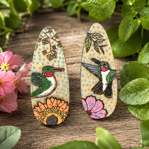 May include: A pair of teardrop-shaped wooden earrings featuring hand-painted hummingbirds, flowers, and green foliage against a light wood background with blue polka dots. The left earring shows a hummingbird near a sunflower, while the right shows a hummingbird in flight near a pink flower.