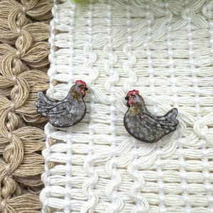May include: Two small, gray chicken-shaped decorative objects with red combs and beaks. The chickens are set against a white and beige woven background. The objects appear to be made of wood or a similar material.