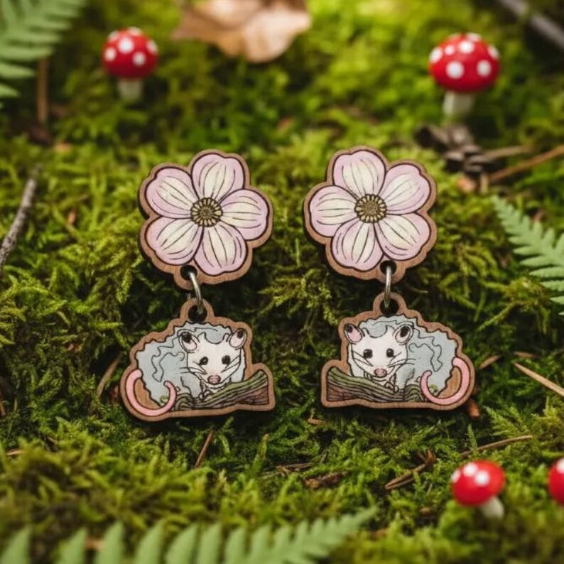 A pair of earrings featuring cartoon opossum characters, surrounded by ferns, mushrooms, and other natural elements, creating a whimsical and nature-inspired scene.