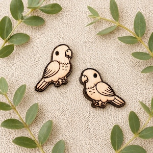 May include: Two wooden bird-shaped embellishments with dark brown outlines and details. The birds are light-colored wood with etched wings, beaks, and feet. Green leaves surround the birds on a neutral background.