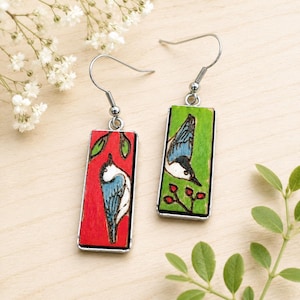 May include: A pair of rectangular drop earrings featuring wood-burned and hand-painted bird illustrations. One earring has a red background with a blue and white bird, while the other has a green background with a blue and white bird and red berries.