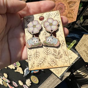 A hand is holding a pair of earrings with a floral design, featuring two pink flowers and a cat-like creature.
