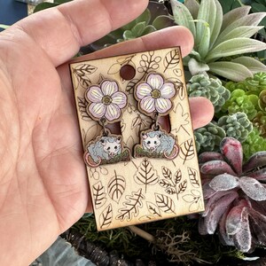 A hand is holding a pair of earrings with a floral design, featuring two opossum-like creatures on the earrings.