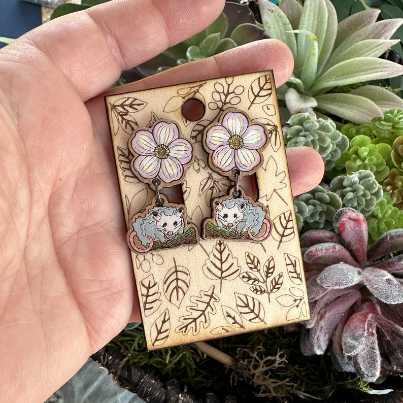 A hand is holding a pair of earrings with a floral design, featuring two opossum-like creatures on the earrings.
