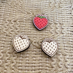 May include: Three wooden strawberry-shaped embellishments. One strawberry is red with a pink outline and green leaves. The other two are natural wood with brown outlines and small dot details, perfect for crafts and DIY projects.