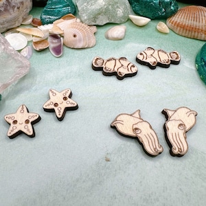 May include: A set of wooden cutouts featuring a variety of sea creatures. The set includes two starfish, two clownfish, and two cuttlefish. The cutouts are all made of natural wood and have a light brown color.
