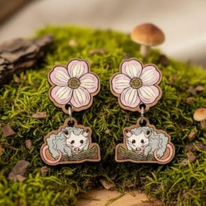 A pair of earrings with a flower design and a small opossum charm, placed on a mossy surface with mushrooms and other natural elements.