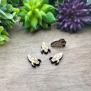 May include: Four wooden rocket ship shaped embellishments. Three are facing forward and one is facing backward. The embellishments are light brown with black outlines.