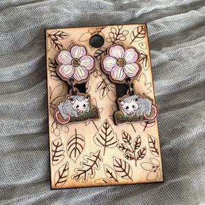 A pair of earrings with a wooden frame and floral designs, featuring two small opossum-like figures.