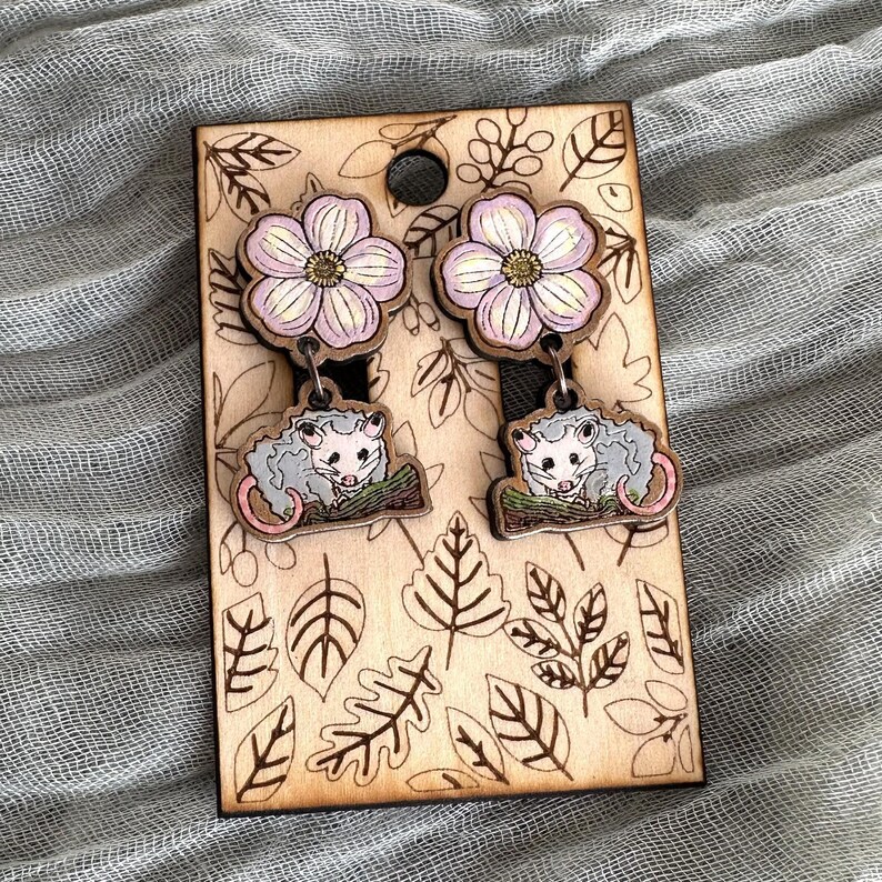 A pair of earrings with a wooden frame and floral designs, featuring two small opossum-like figures.