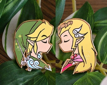 May include: Two gold-colored enamel pins depicting cartoon characters from the Legend of Zelda video game series. The characters are Link and Zelda, and they are facing each other with their eyes closed. Link is wearing his signature green tunic and hat, while Zelda is wearing a pink dress with a gold trim.