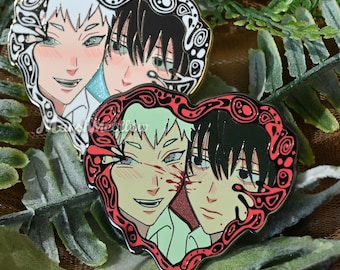 May include: Two heart-shaped enamel pins featuring anime-style portraits. One pin has a white and black design, the other a red and black design. The portraits depict two characters each, with detailed facial features and expressions.