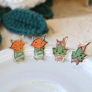 May include: Four enamel pins featuring cartoon characters with autumnal leaves. The characters are green, orange, and pink with green leaves. The pins are on a white surface.