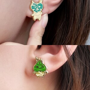 May include: Gold-tone earrings featuring a cartoon character with a green leaf on its head. The character is wearing a white and brown striped shirt.