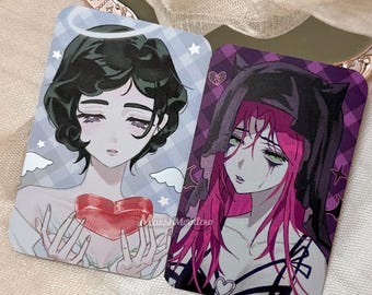 Puede incluir: Dos tarjetas coleccionables rectangulares con ilustraciones estilo anime. La tarjeta izquierda muestra una figura con cabello oscuro, un halo y un corazón rojo. La tarjeta derecha representa una figura con cabello rosa y capucha negra.