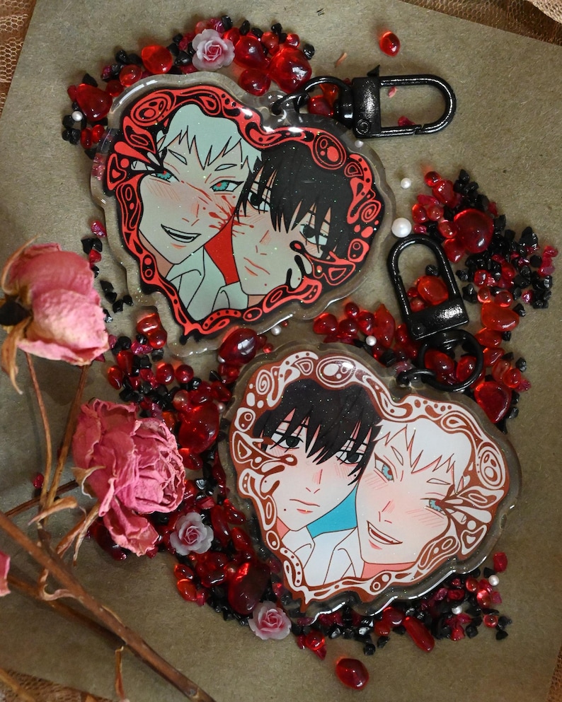 K&ouml;nnte beinhalten: Herzf&ouml;rmige Schl&uuml;sselanh&auml;nger mit Anime-Illustrationen von zwei sich umarmenden Charakteren. Die Schl&uuml;sselanh&auml;nger haben einen dekorativen roten Rand und sind von roten und schwarzen Perlen, kleinen Rosen und Trockenblumen umgeben. Jeder Schl&uuml;sselanh&auml;nger hat einen schwarzen Verschluss.