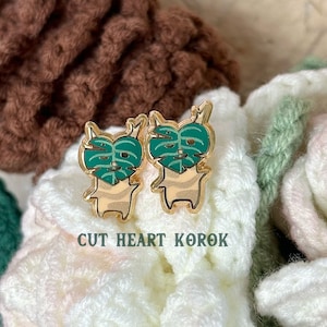 May include: Two gold-colored pins featuring a cartoon character with a green monstera leaf covering its face. The pins are on a white and brown crocheted surface. The text "CUT HEART KOROK" is below the pins.