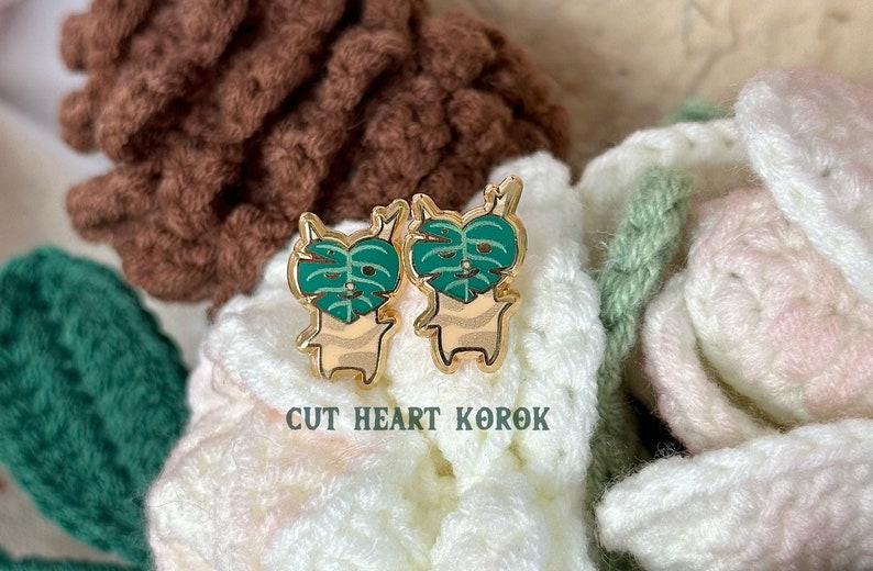May include: Two gold-colored pins featuring a cartoon character with a green monstera leaf covering its face. The pins are on a white and brown crocheted surface. The text "CUT HEART KOROK" is below the pins.