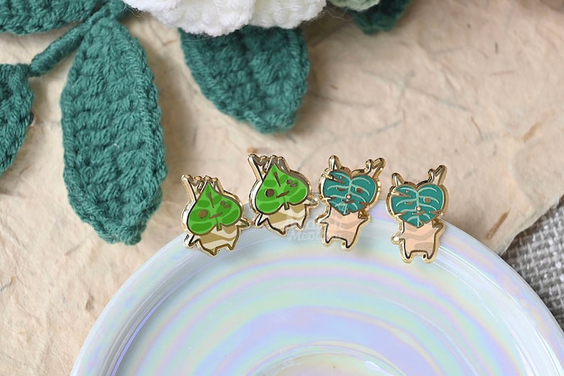 May include: Four gold-colored enamel pins featuring a cartoon character with a green leaf on its head. The character has a light brown body and a white face with black eyes. The pins are arranged in a row on a white iridescent surface.