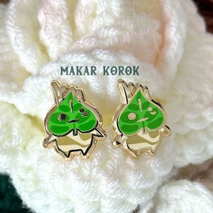 May include: A pair of gold-colored earrings featuring a green leaf design with a smiling face. The earrings are shaped like a small, cartoon-like character with a green leaf for a head and a white body. The text "MAKAR KOROK" is visible in the background.