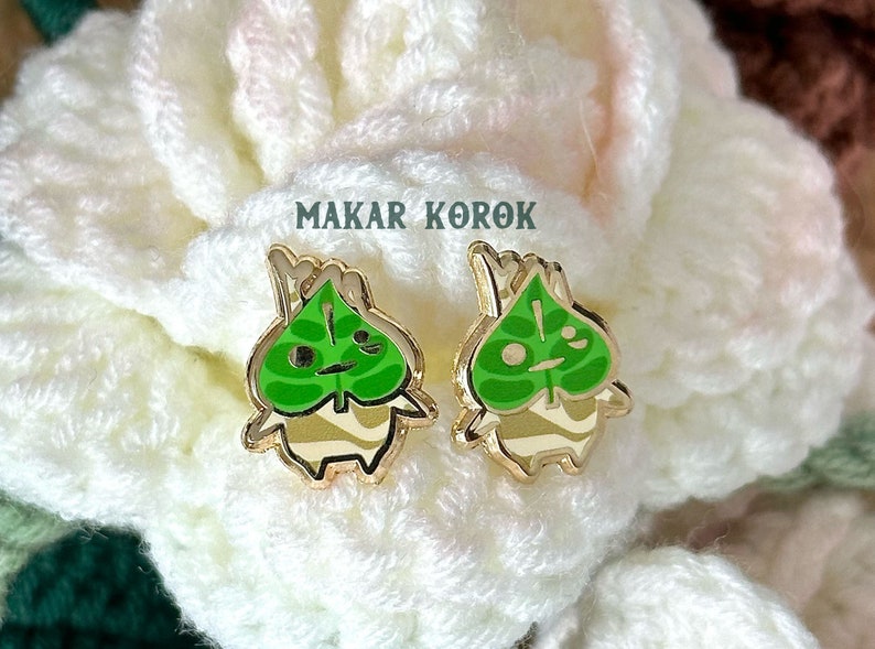 May include: A pair of gold-colored earrings featuring a green leaf design with a smiling face. The earrings are shaped like a small, cartoon-like character with a green leaf for a head and a white body. The text "MAKAR KOROK" is visible in the background.